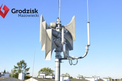 Syreny powiadamiania alarmowego zamontowane na dachu budynku, w tle widoczne dachy innych budynków oraz niebieskie niebo. W lewym górnym rogu widoczny logotyp Gminy Grodziska Mazowiecki