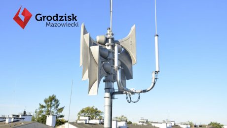 Syreny powiadamiania alarmowego zamontowane na dachu budynku, w tle widoczne dachy innych budynków oraz niebieskie niebo. W lewym górnym rogu widoczny logotyp Gminy Grodziska Mazowiecki